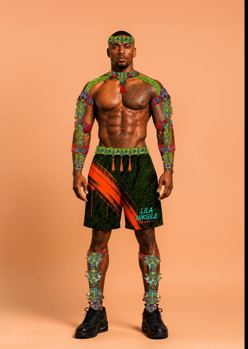 A muscular male model stands against a solid peach studio background wearing a matching multicolor tribal-patterned outfit including a headband, armbands, shorts, and leg wraps, along with black sneakers. The shorts feature green, orange, and black patterns with 'LIL' JAZZ' printed on the side.