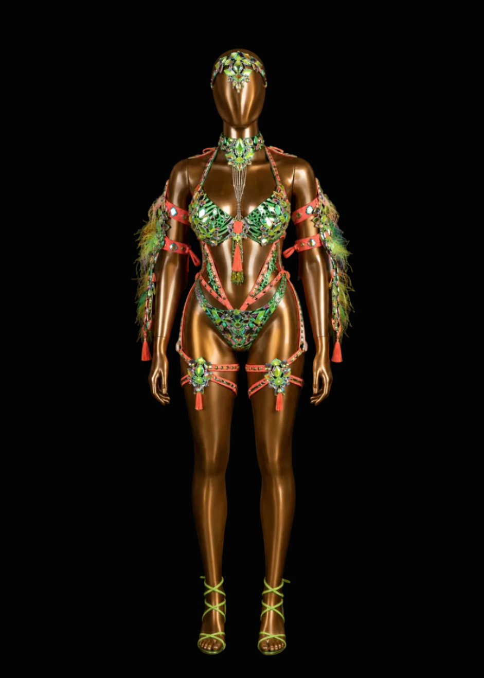 A metallic bronze mannequin stands against a black background wearing an ornate green and red embellished lingerie-style costume with feathered shoulder details, matching headpiece, and strappy high-heeled sandals.
