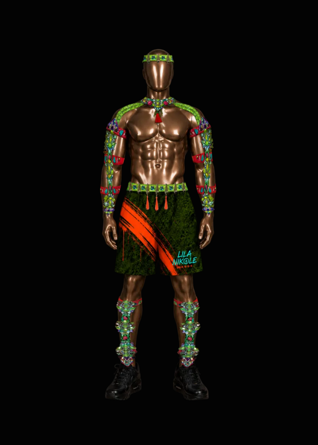 A metallic bronze mannequin wearing a highly decorated tribal-inspired outfit with green patterned arm and leg wraps, a green skirt with orange diagonal stripes, a red chest piece, and a matching headband, set against a solid black background.