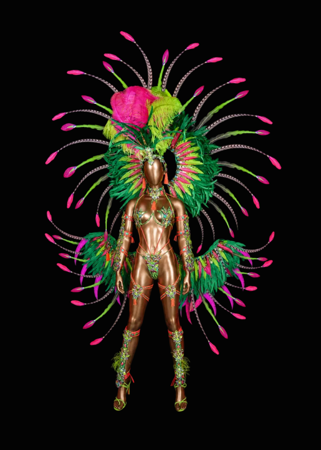 A metallic bronze mannequin wearing an elaborate carnival costume with a large feathered headdress and matching feathered shoulder pieces in green, pink, and yellow, set against a solid black background.