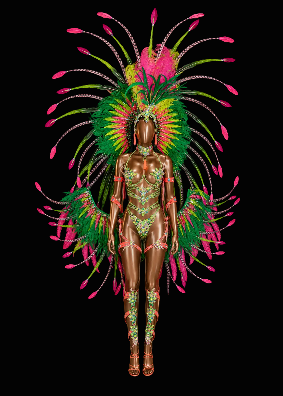 A full-body mannequin wearing an elaborate carnival-style costume with a large feathered headdress and matching feathered shoulder/back pieces, shown against a solid black background. The mannequin has a metallic bronze finish, and the costume features bright green, pink, yellow, and orange feathers with red trim.
