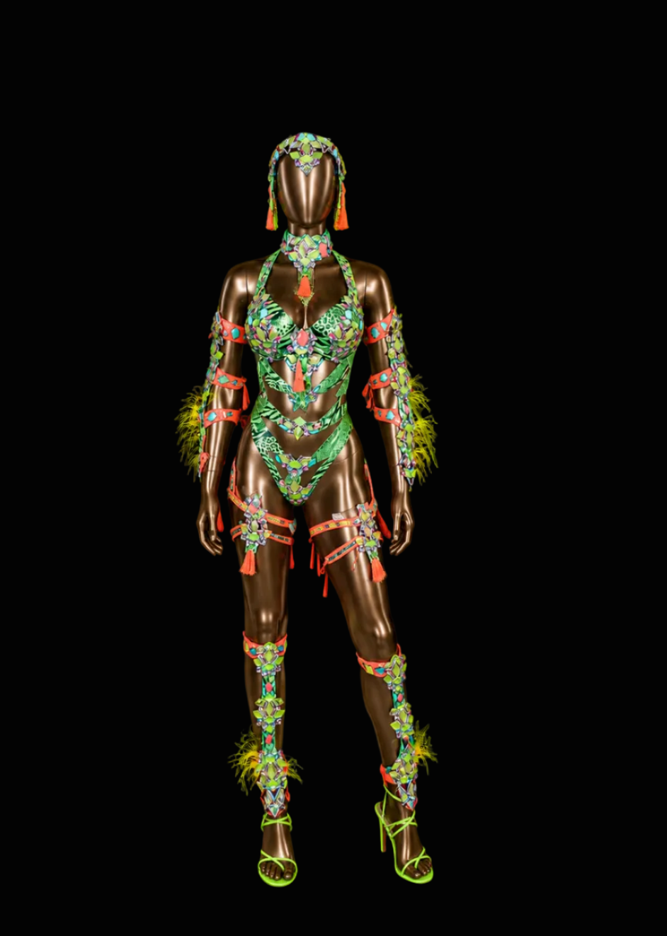 A metallic bronze mannequin wearing an ornate, highly embellished green and multicolor costume with feathered accents, set against a solid black background.
