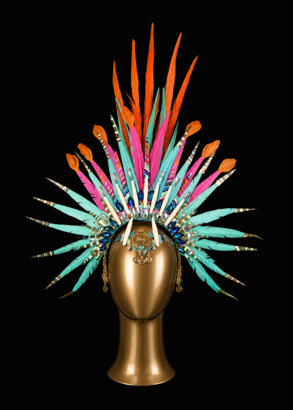 A vibrant, multicolored feather headdress displayed on a metallic gold mannequin head against a solid black background. The headdress features a dense array of feathers in bright turquoise, orange, pink, and white, with gold-toned metal accents and decorative details.