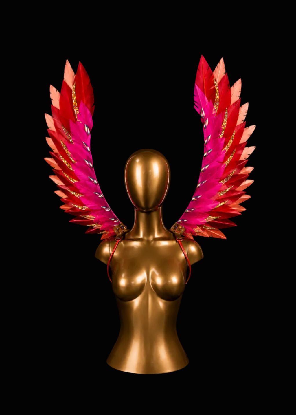 A pair of large, vibrant feather wings in shades of pink, red, and orange with gold glitter accents, displayed on a metallic gold mannequin bust against a solid black background.