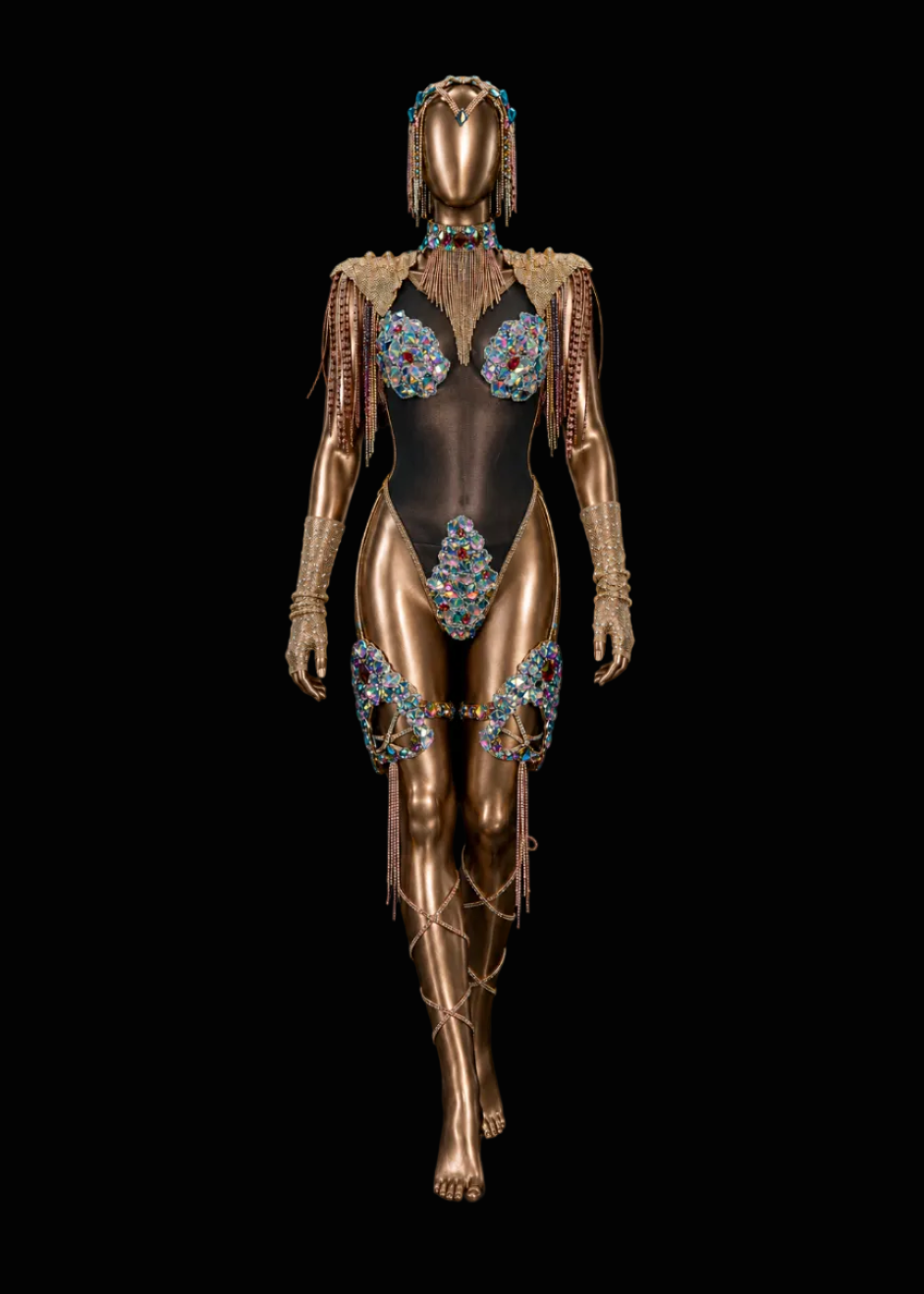 A metallic gold mannequin styled as a model, wearing a black bodysuit heavily embellished with blue, red, and green rhinestones, along with matching rhinestone arm cuffs and leg wraps, set against a solid black background.