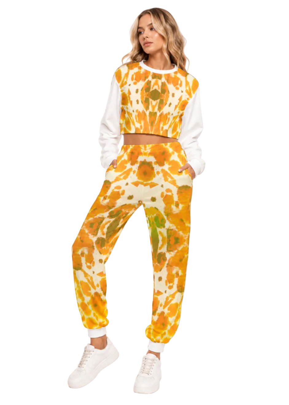 A woman modeling a matching two-piece lounge set featuring a white long-sleeve crop top and jogger pants with a vibrant orange, yellow, and green tie-dye pattern, paired with white sneakers on a white studio background.