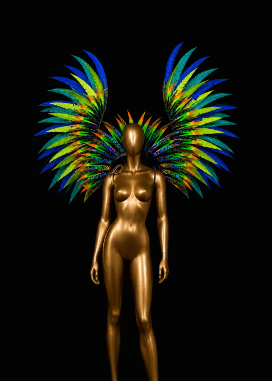 A glossy metallic gold female mannequin stands against a solid black background, wearing large feathered wings in bright blue, green, yellow, and orange.
